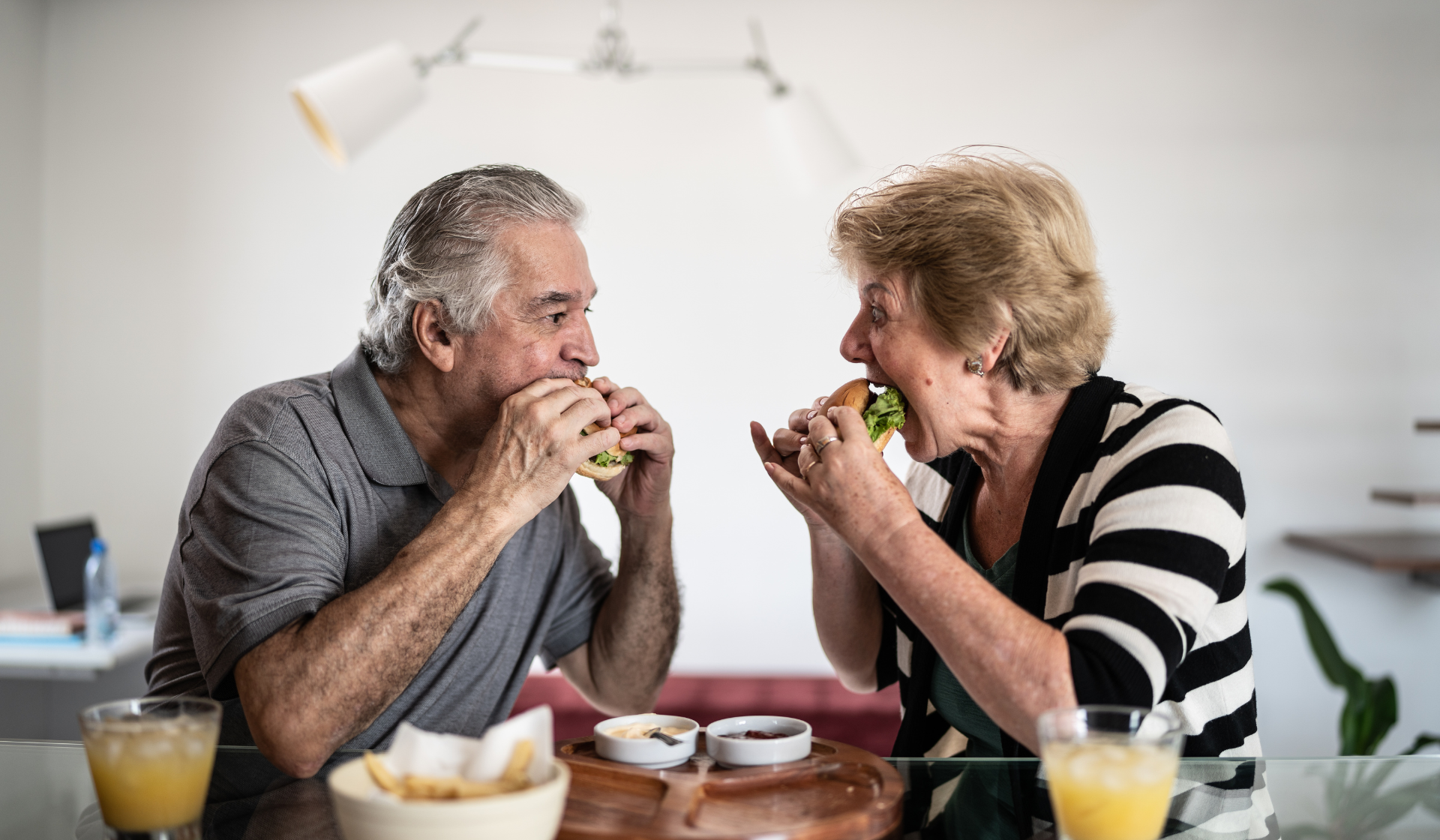 old man and old lady looking at each other as they bite into a burger